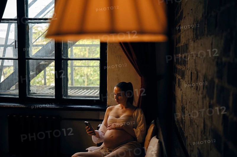 In a cozy room filled with natural light, a pregnant woman sits comfortably on sofa and making delivery order on mobile app