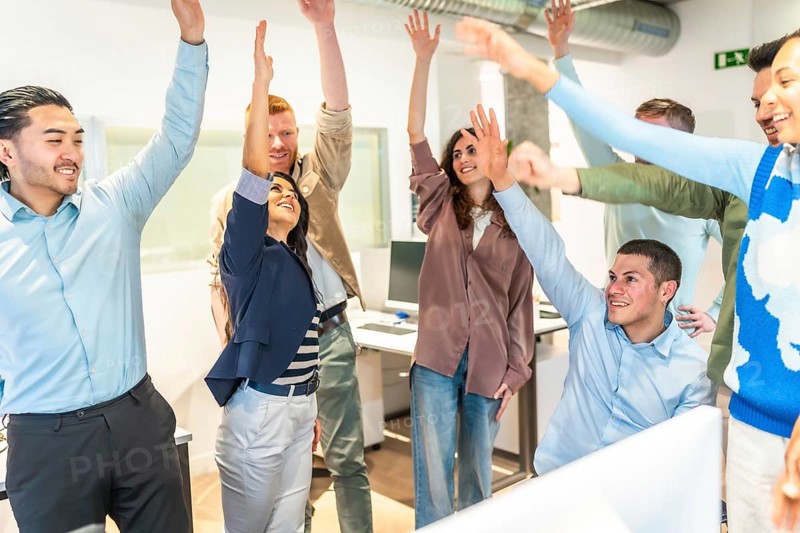 Multicultural businesspeople celebrating success in a bright office, raising arms in unity, showcasing teamwork and enthusiasm for achievement