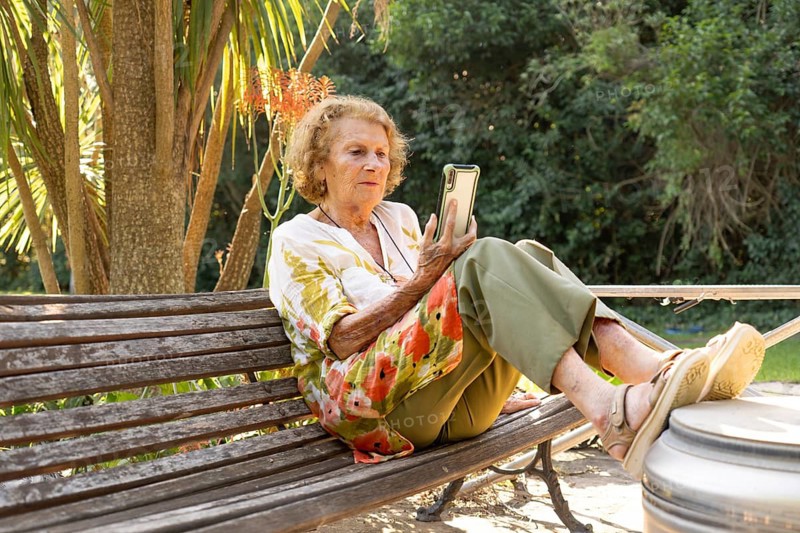 Senior woman enjoying leisure time outdoors, sitting comfortably on a wooden bench in a lush park, using her smartphone for communication or entertainment