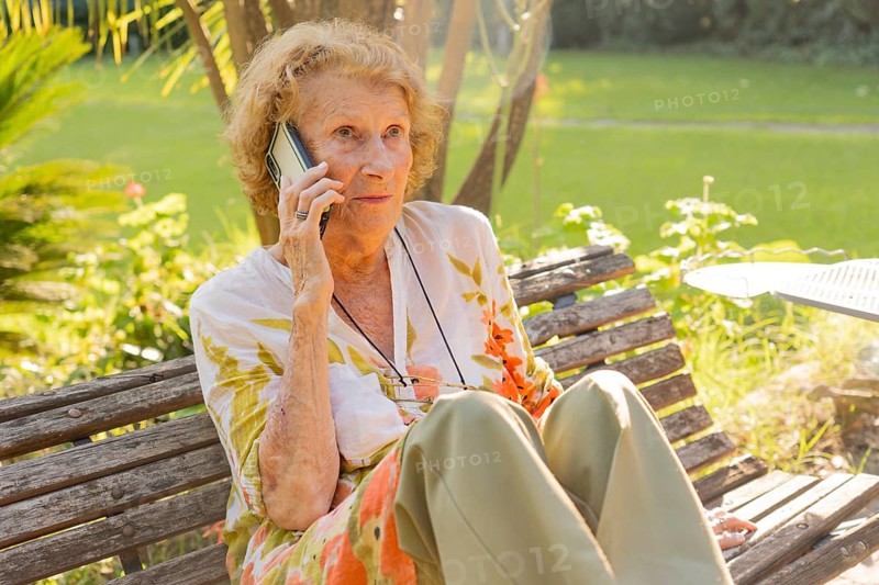 Senior woman enjoying a conversation on her mobile phone while sitting on a wooden bench in a serene park setting, embracing technology and staying connected in her retirement