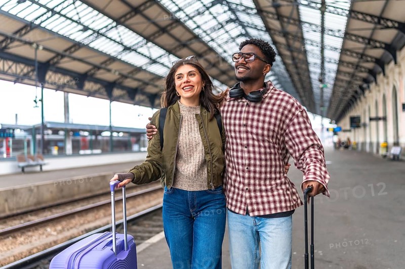 Young multi ethnic couple walking on a platform at the train station, smiling and holding suitcases, ready to start their vacation