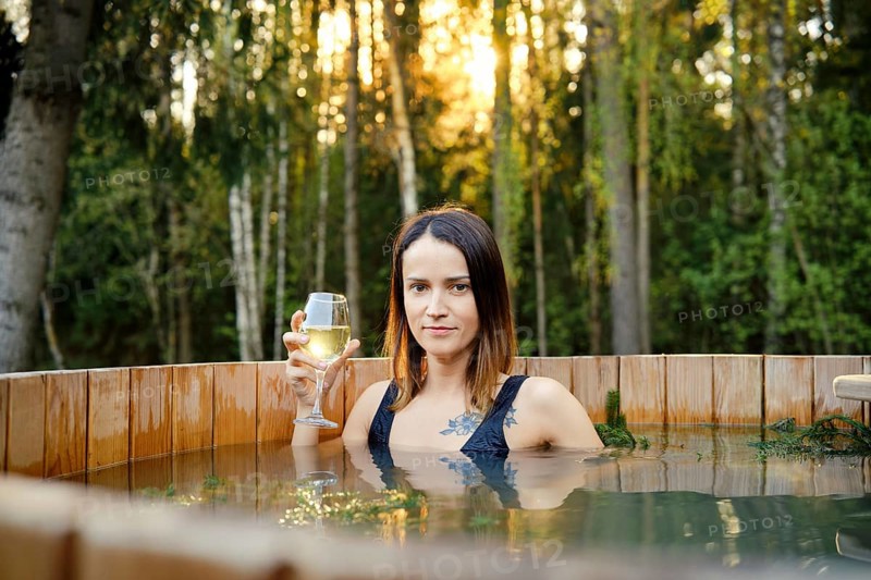 A woman with long hair holds a glass of wine while relaxing in a wooden hot tub. The sun sets behind her, casting a warm glow and highlighting the lush greenery surrounding the serene forest setting