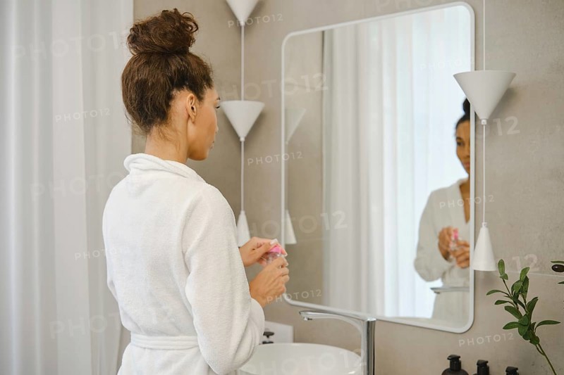 A woman wearing a white robe stands in front of a large mirror in a stylish bathroom. She opens a jar of micellar water and looks at her reflection