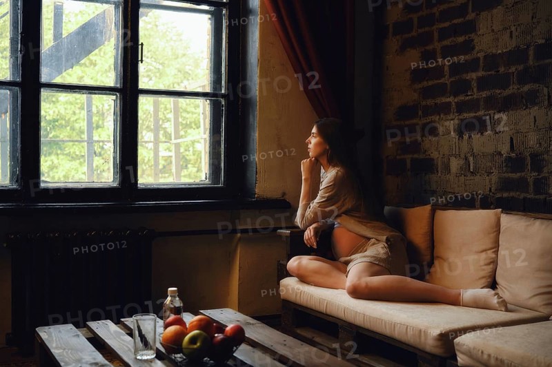 A pregnant woman sits comfortably on a couch, gazing thoughtfully out the window. She enjoys a quiet moment in her cozy home, surrounded by natural light and a peaceful atmosphere