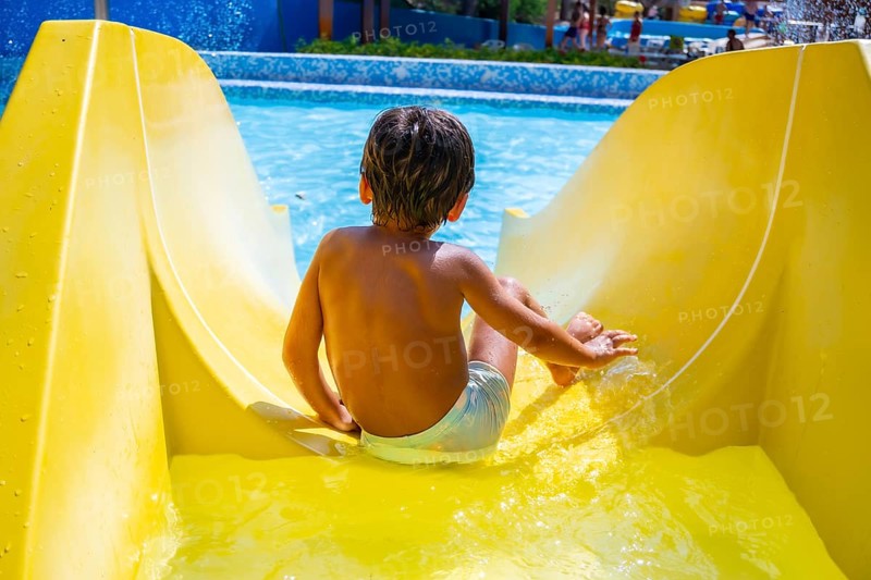 Young boy enjoying summer holidays sliding down yellow waterslide into swimming pool in aqua park