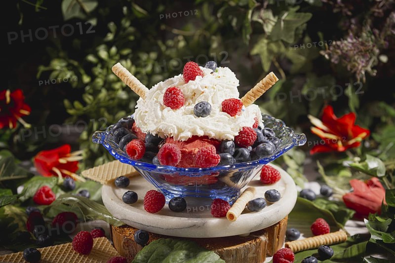 Colourful sundae with raspberries and blueberries, decorated with cream and waffle, surrounded by leaves and flowers