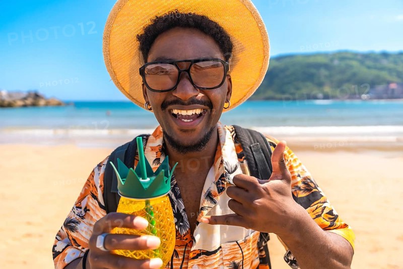 Young man enjoying summer vacation on a tropical beach, holding a pineapple shaped cocktail and gesturing the shaka sign