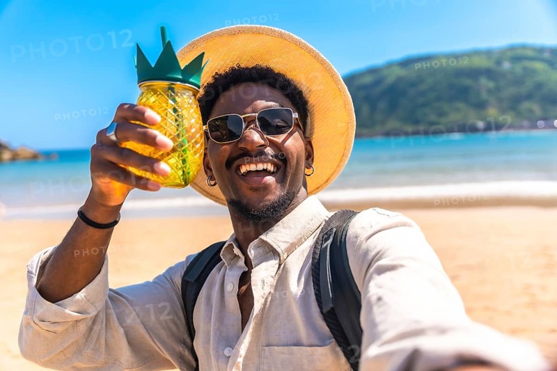 Young man wearing sunglasses and straw hat holding pineapple shaped cocktail while taking selfie on beach