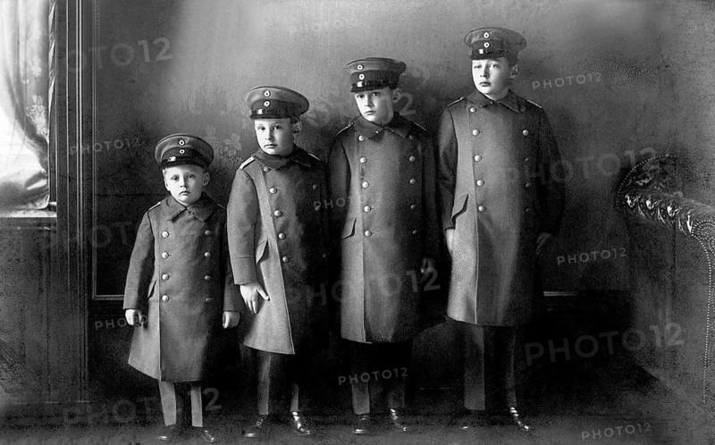 Four uniformed boys lined up in height, military, militarism, 1910s, group of boys in uniforms and caps, posing in front of a neutral backdrop, serious facial expression, historical photo