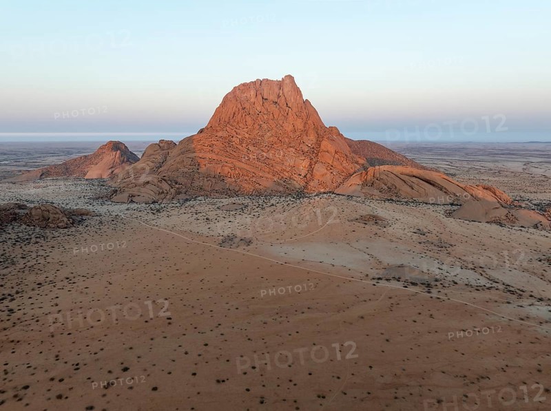 The isolated Spitzkoppe mountain (1728 m) majestically rises above the surrounding desert plains. In the distance the coast and the Atlantic Ocean. At dawn. Aerial view. Drone shot. Damaraland, Namibia
