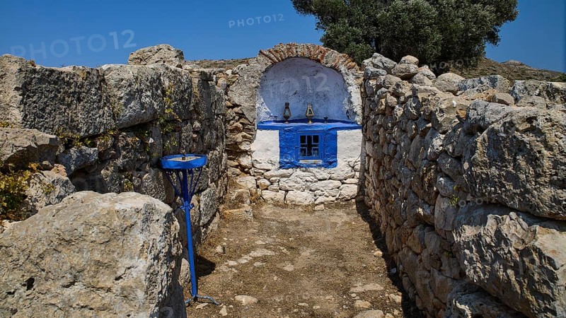 Small stone chapel with blue façade embedded in ancient ruins under a bright blue sky, Artemis Temple, Artemis, Diana, goddess of the hunt, altar. Church, tower, near the airport in the north of the island, Leros, Dodecanese, Greek Islands, Greece