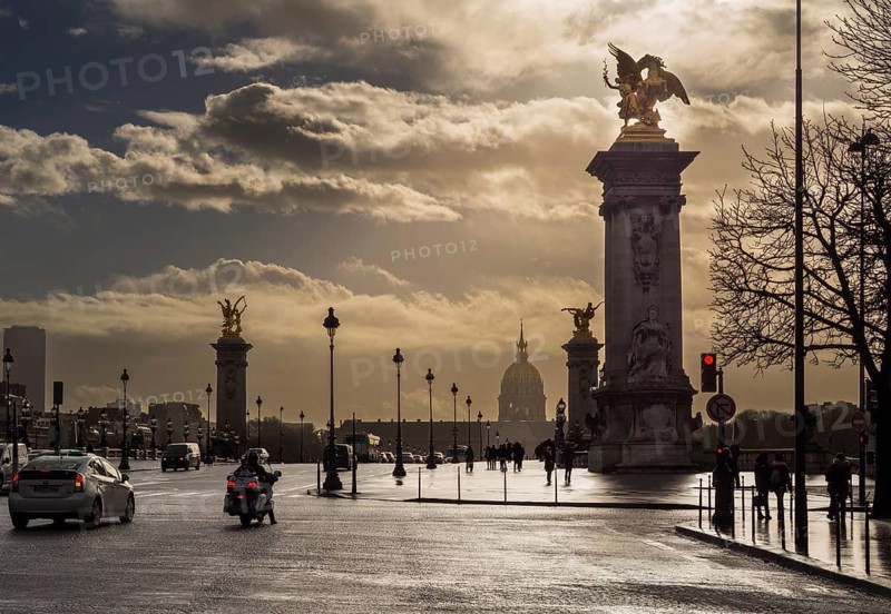 Clouds gather above the iconic Alexandre III Bridge as vehicles and people navigate the bustling streets of Paris. The scene captures the city's timeless charm and vibrant energy, France