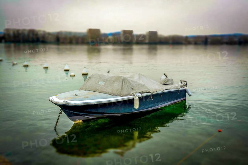 A solitary boat sits anchored in still waters, surrounded by gentle mist. The shoreline is faintly visible in the background, creating a serene atmosphere at dawn, Annecy, Haute Savoie, Auvergne Rhone Alpes, France