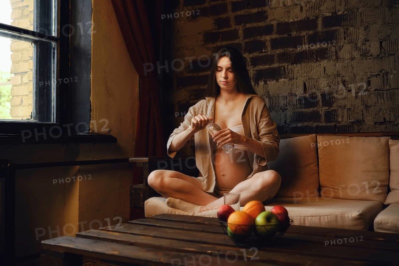 A pregnant woman sits comfortably on a sofa in her home, enjoying a moment of calm. She is focused on opening a bottle with water