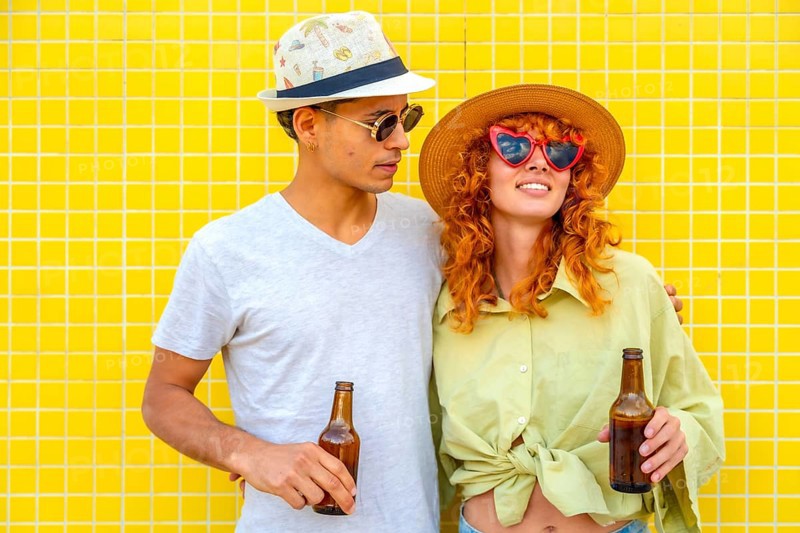 Young couple wearing hats and sunglasses, holding beer bottles and smiling joyfully against a vibrant yellow tiled wall, radiating happiness
