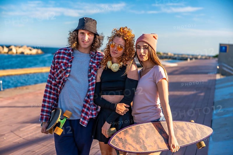Three friends with skateboards relax by the seaside, enjoying the sunny weather and casual outdoor vibes on a wooden boardwalk