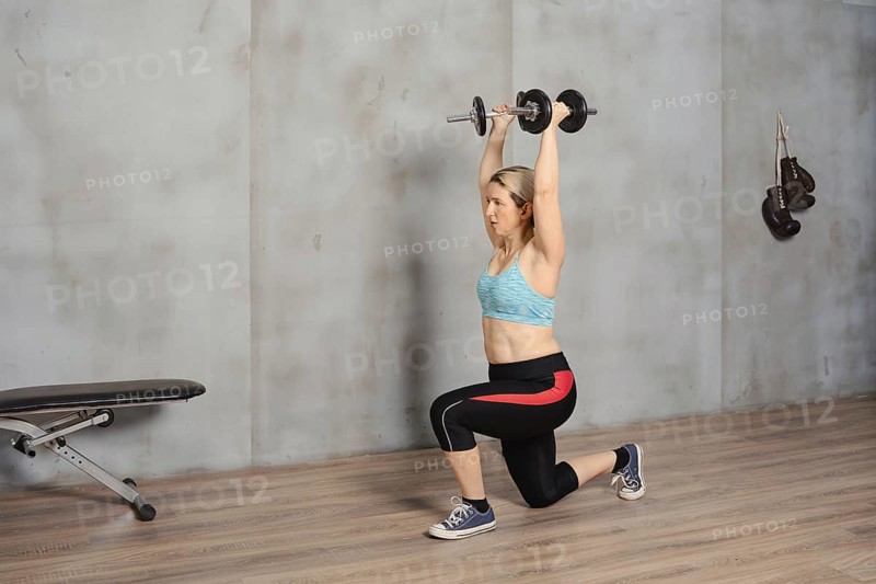 A woman is engaged in a strength training workout, lifting dumbbells overhead while in a lunge position. She is inside a gym, wearing athletic clothing, focusing on her fitness routine