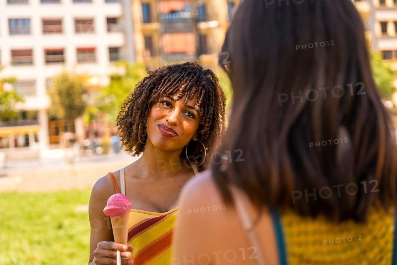 Young woman enjoying a strawberry ice cream cone while talking to her friend in a city park on a summer day