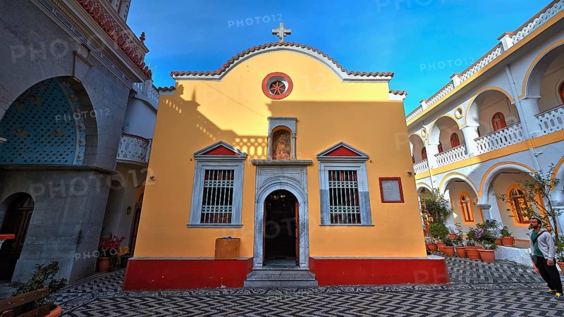 Yellow church with courtyard, cross and baroque elements, surrounded by plants, Holy Week, Bay of Panormitis, Panormitis Monastery, Symi, Dodecanese, Greek Islands, Greece