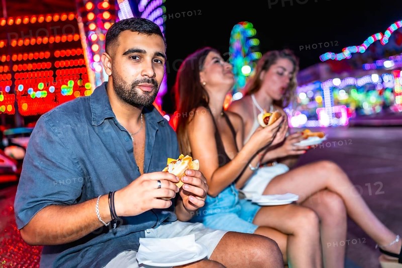 Young adults savoring delicious hot dogs while enjoying the vibrant atmosphere of an amusement park at night, surrounded by colorful lights