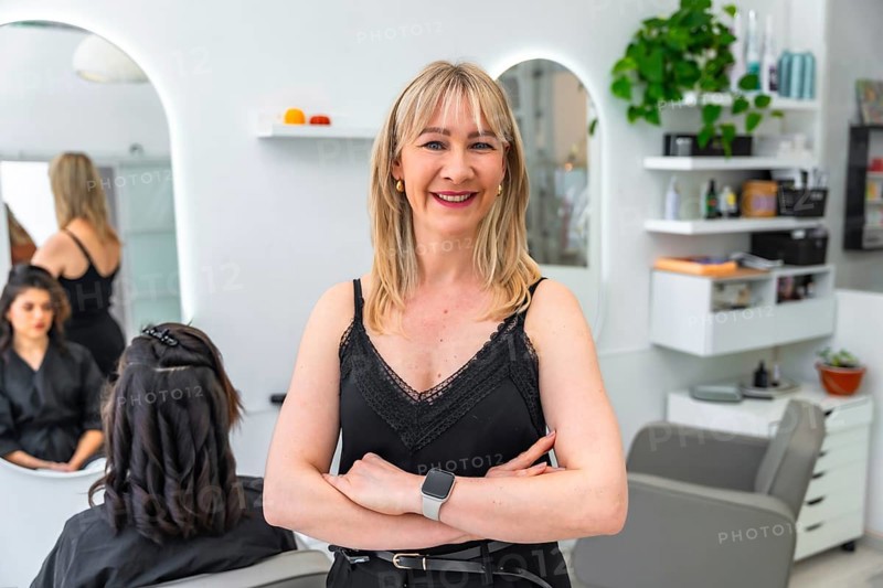 Confident female entrepreneur and owner of a beauty salon smiles with arms crossed as her employee styles a client's hair