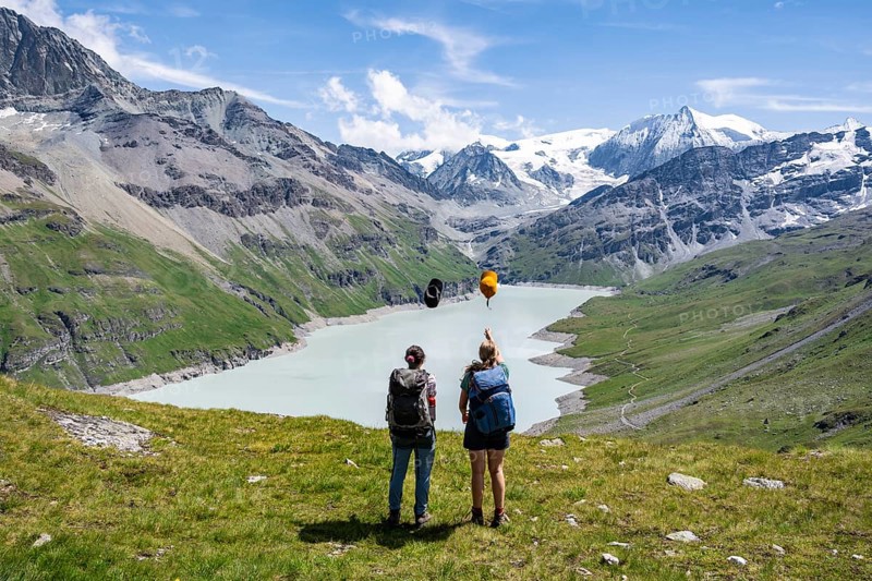 Two female friends throw their hats in the air, female mountaineers in front of a blue mountain lake, Lac des Dix reservoir, in the background mountain peak Mont Blanc de Cheilon with glacier, Hérménence, Valais Alps, Valais, Switzerland