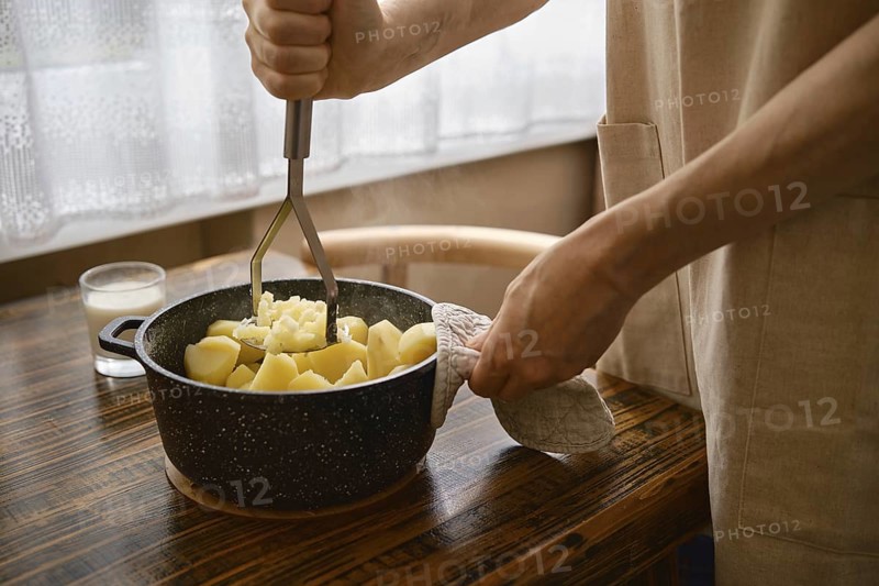 A person is mashing boiled potatoes in a pot. His hand grips a masher while holding a pot with another hand