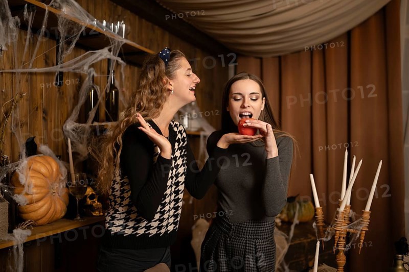 Two friends are playfully engaging in a Halloween celebration, surrounded by festive decorations. One friend holds a red apple while the other smiles and gestures in a cozy, spooky atmosphere