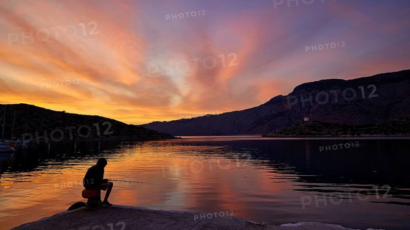 Angler sitting on the lakeshore during a colourful sunset against a mountainous backdrop, Holy Week, Bay of Panormitis, Panormitis Monastery, Symi, Dodecanese, Greek Islands, Greece
