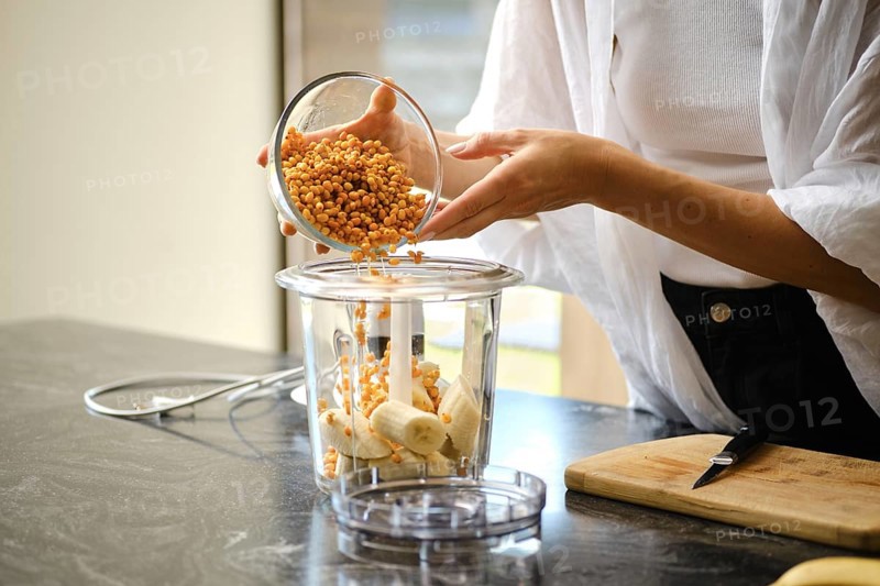 A person carefully adds sea buckthorn berries to a blender containing sliced bananas. The setting is a modern kitchen with natural light, highlighting the vibrant colors of the ingredients