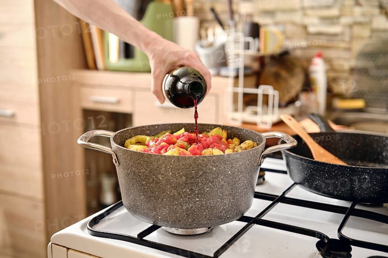 A person pours red wine into a pot filled with pork and vegetables making aromatic sauce