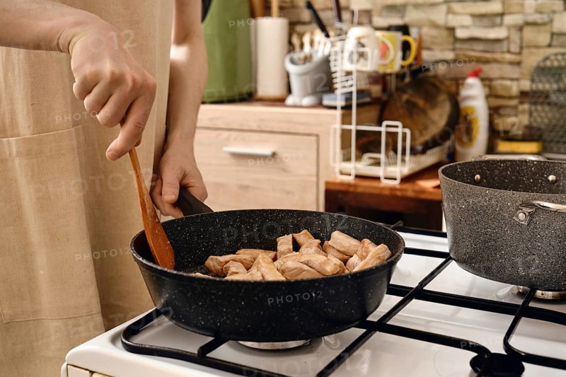 A person stirs diced pork meat in a frying pan on a gas stove. The kitchen is modern, featuring stone walls and organized utensils