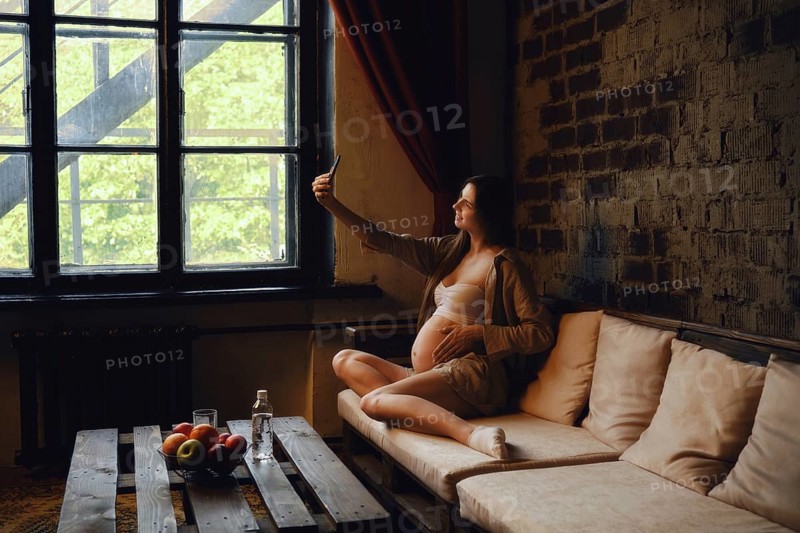 Relaxing at home, a pregnant woman sits on a cozy couch, taking a selfie and posting it in her social media. She is happy to share her peaceful moment