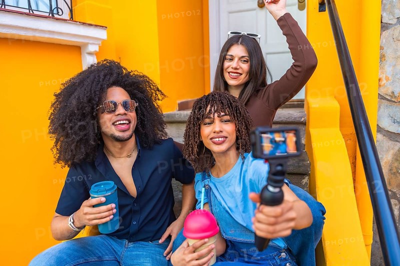 Three happy multi ethnic content creators filming a video blog with a gimbal and drinking smoothies on colorful steps