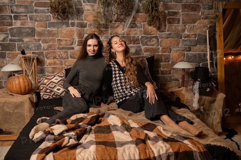 Two women relax together on a bed surrounded by autumn decor, including pumpkins and warm blankets, in a rustic setting adorned with stone walls and soft lighting