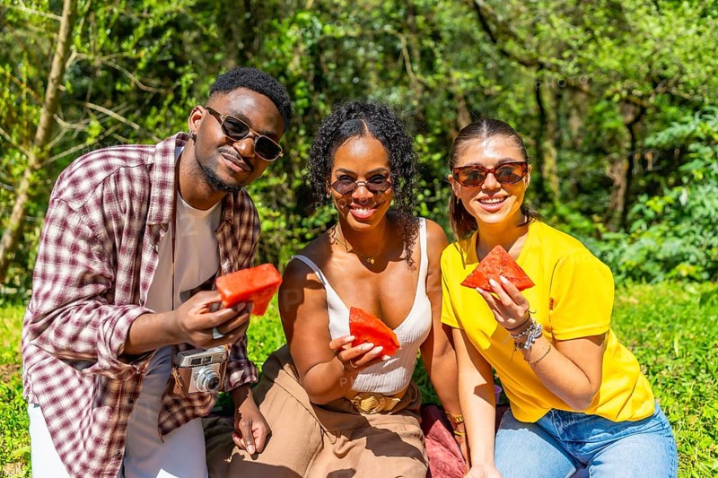 Three friends are sharing watermelon slices during a summer picnic in a lush park, enjoying the sunny weather and each other's company
