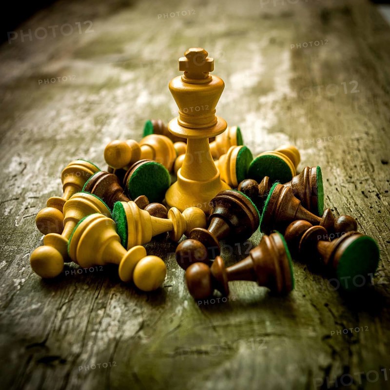 Chess pieces are strewn across a rustic wooden table, with a prominent king piece standing tall amidst the fallen soldiers, indicating a moment of strategic conflict