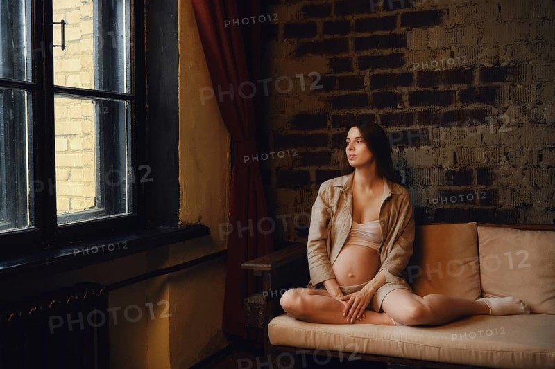 A relaxed pregnant woman sits on a couch in her home, gazing thoughtfully out the window. Soft light filters through, highlighting her gentle pose and the warmth of the interior