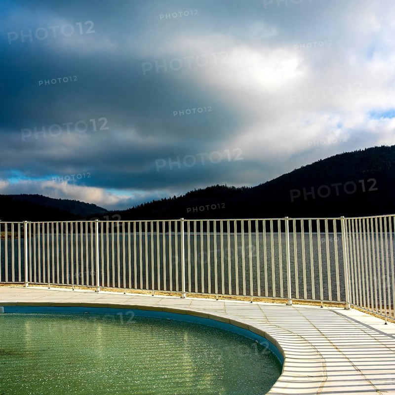 A serene lake reflects the overcast sky, with gentle ripples on the surface. Fencing outlines the pool area, while rolling hills provide a tranquil backdrop, Auvergne Rhone Alpes, France