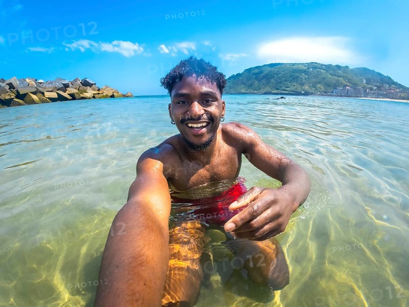 Young man enjoying a sunny summer day, capturing a joyful selfie while bathing in crystal clear sea water, surrounded by vibrant nature