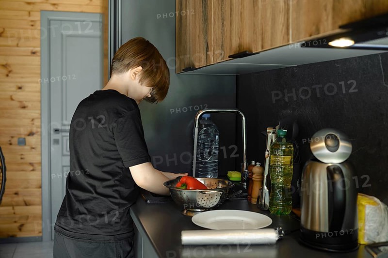 A boy washes vegetables in the sink and puts them in a colander, standing in profile at a sleek kitchen counter