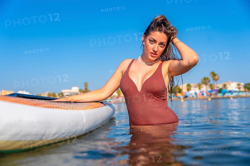 Young woman wearing swimsuit posing in the sea next to her paddle board, enjoying summer vacation on a sunny day
