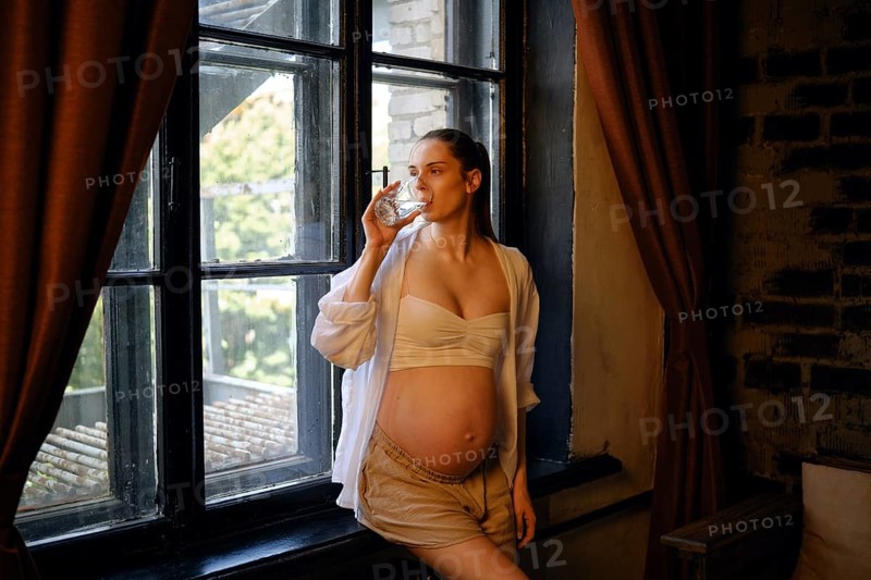 A pregnant woman stands by a large window at home, enjoying a drink from a glass. She wears a comfortable outfit and looks relaxed as natural light illuminates the cozy space