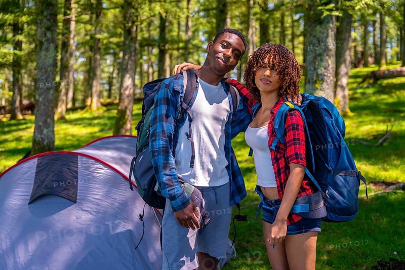 Two hikers with backpacks are posing next to their tent, enjoying the beautiful forest scenery during a camping trip