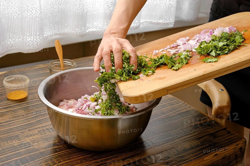A hand adds chopped cilantro and onions into a silver mixing bowl with minced meat on a wooden table. The soft light from the window highlights the fresh ingredients