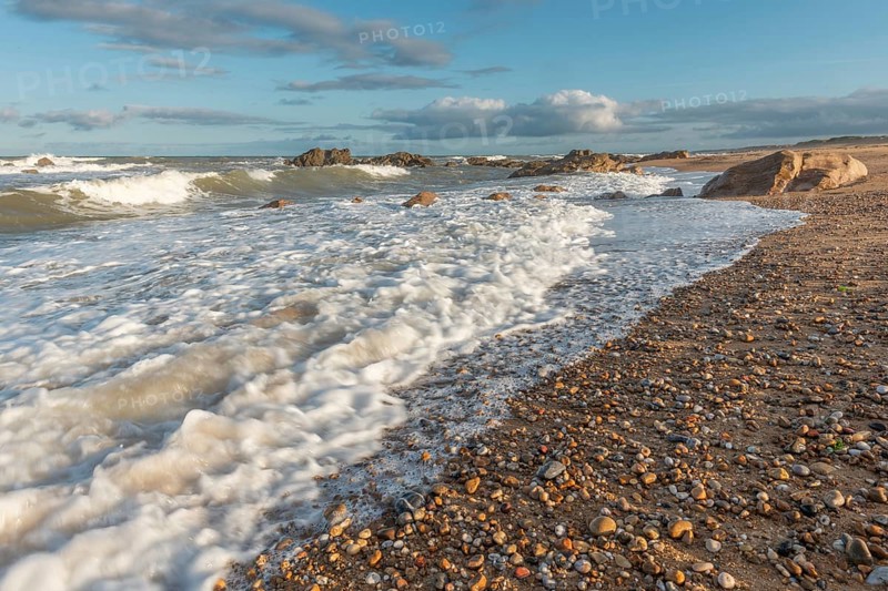 The waves crash gently against a rocky shoreline at sunset, creating a tranquil atmosphere. The sandy beach is strewn with pebbles and surrounded by a beautiful sky. Plage de l'Aubraie, Les Sables d'Olonne, Vendee, France