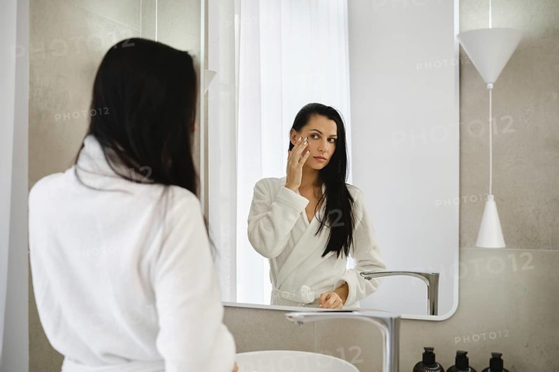 In a bright bathroom, a woman dressed in a white robe applies skincare to her face while looking into a mirror. Natural light fills the space, creating a serene atmosphere for self-care