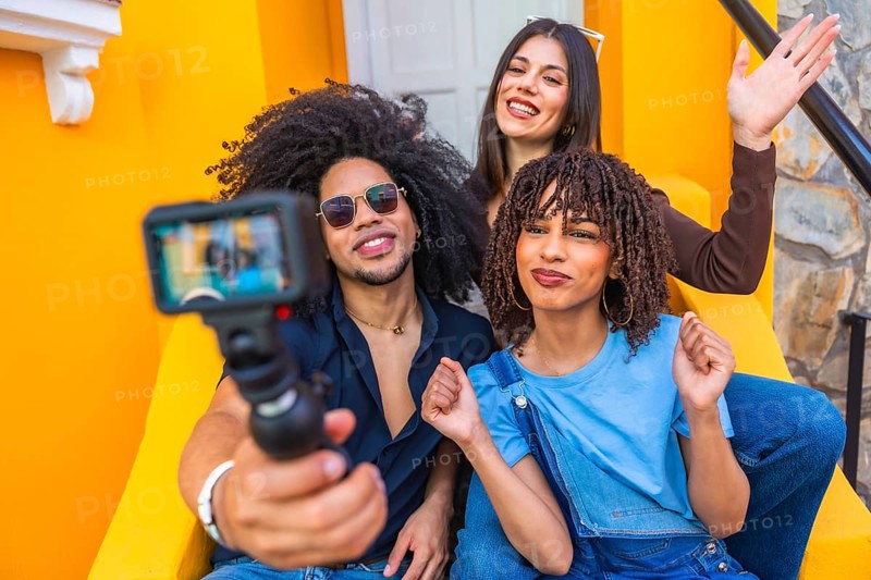 Three happy multi ethnic influencers sitting on yellow steps are filming a vlog with an action camera