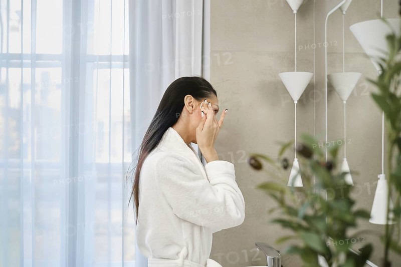 A woman in a white bathrobe is elegantly applying skincare in a contemporary bathroom. Soft natural light filters through sheer curtains, enhancing the calming atmosphere and minimalist design