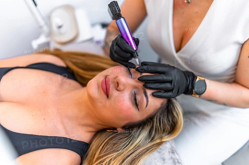 Beautician wearing black gloves using a dermopigmentation machine applying permanent eyeliner makeup on a client lying on a stretcher